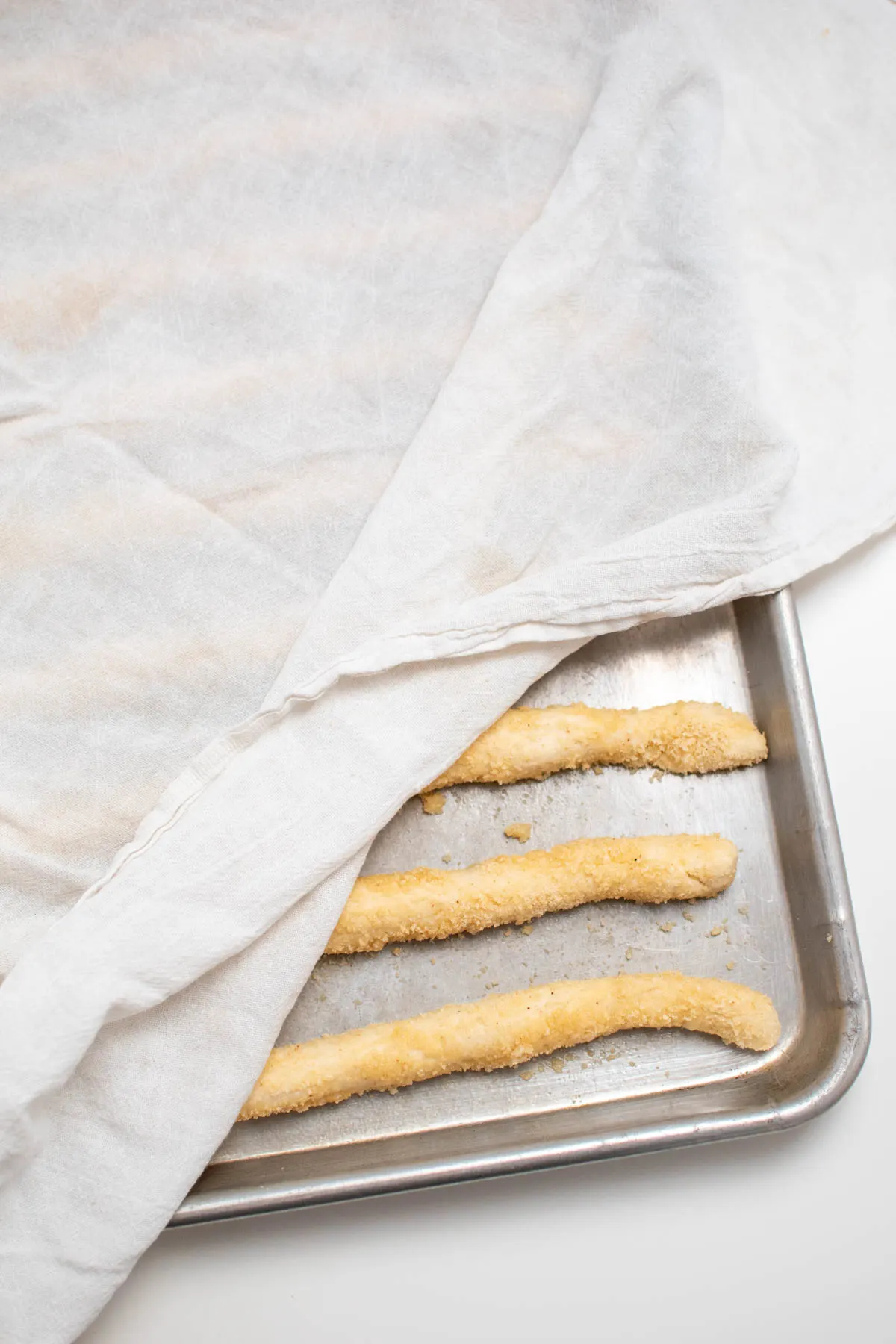 Uncooked breadsticks on baking pan partially covered with white tea towel.