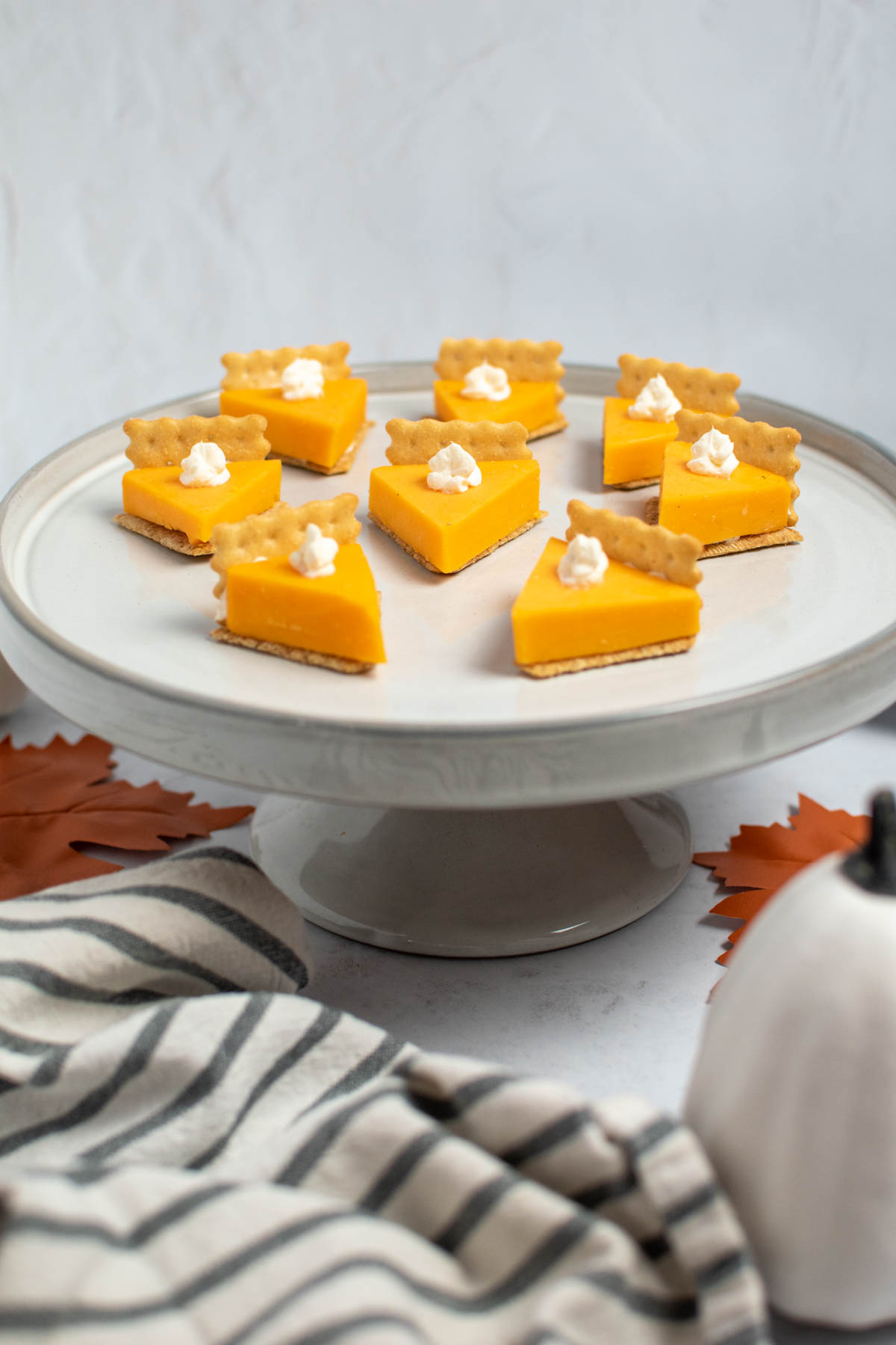 Cheese and cracker pumpkin pie bites on gray cake stand surrounded by towel and pumpkin.