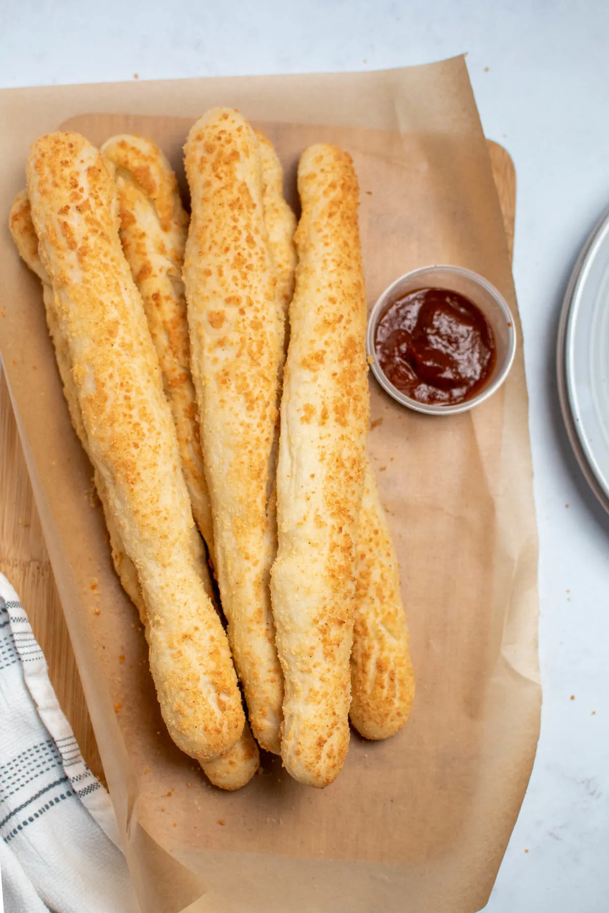 Several parmesan cheese breadsticks on parchment paper with small cup of marinara sauce nearby.