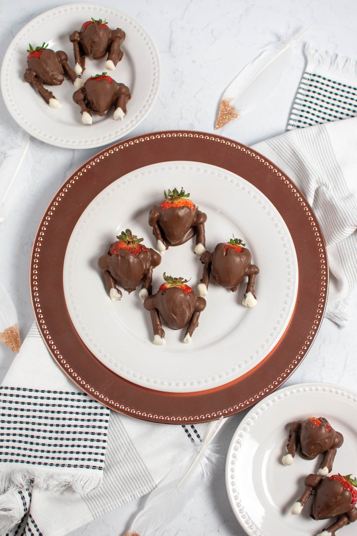 Chocolate dipped strawberry turkeys on white plates with feathers and kitchen towel all on table.