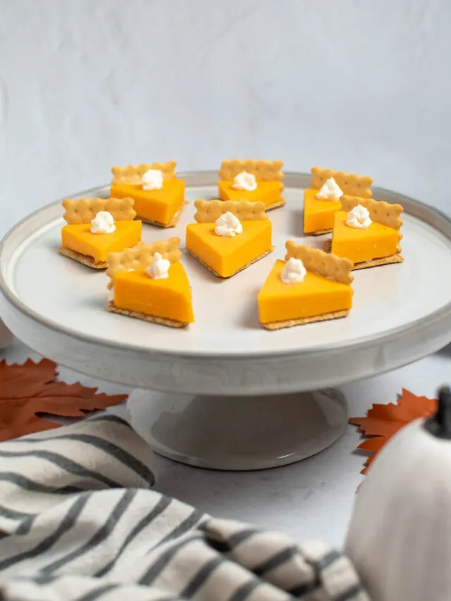 Cheese and cracker pumpkin pie bites on gray cake stand surrounded by towel and pumpkin.