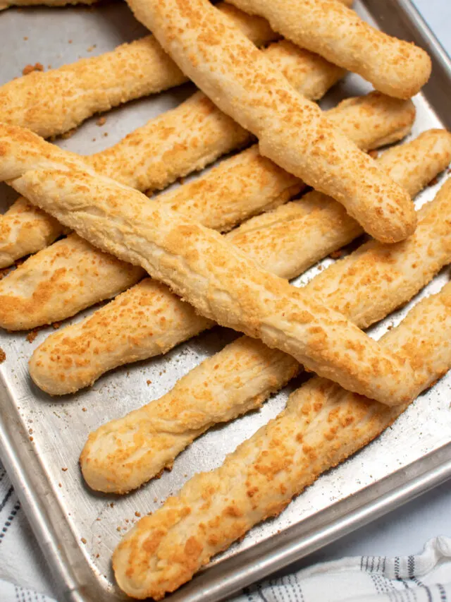 Several Rhodes Rolls breadsticks with parmesan stacked on metal baking sheet.