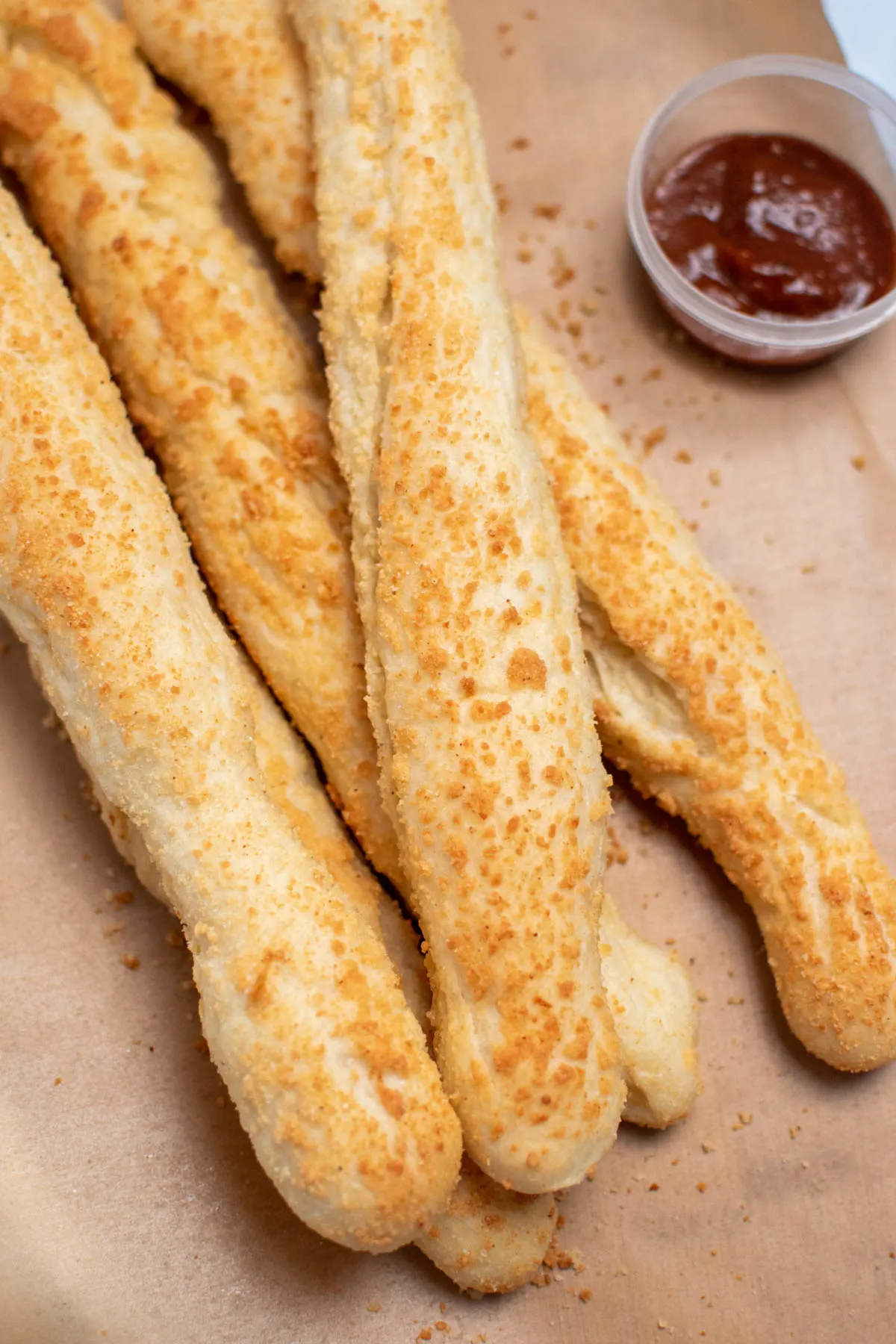 Several parmesan garlic breadsticks stacked on parchment paper with small cup of marinara sauce nearby.