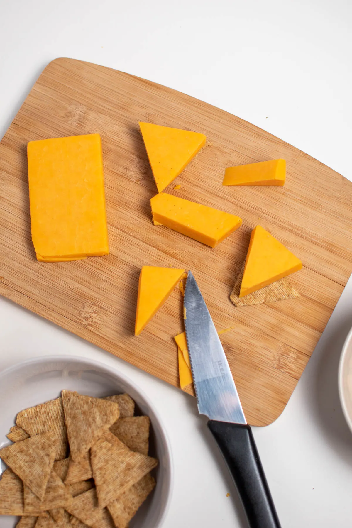 Triangle pieces of cheese on wood cutting board with knife and bowl of crackers nearby.