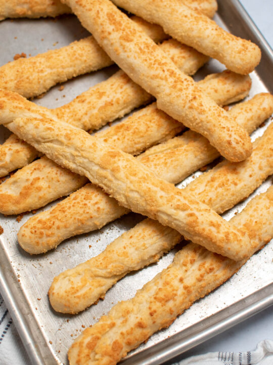 Several Rhodes Rolls breadsticks with parmesan stacked on metal baking sheet.
