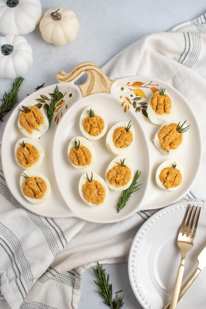 Thanksgiving deviled eggs on pumpkin-shaped platter with fake pumpkins and kitchen towel nearby.