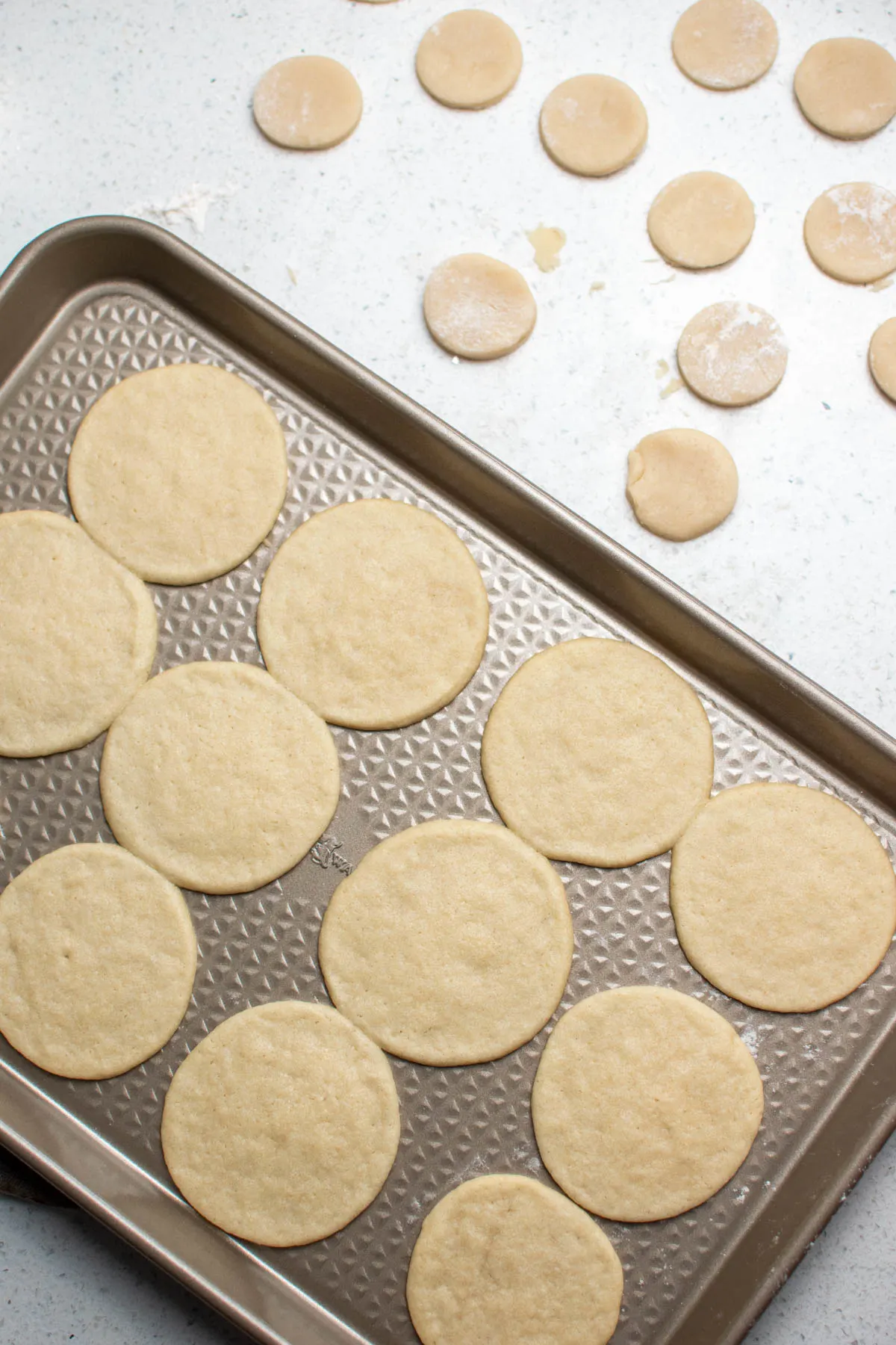 Cooked sugar cookies on baking sheet next to small dough circles on countertop.