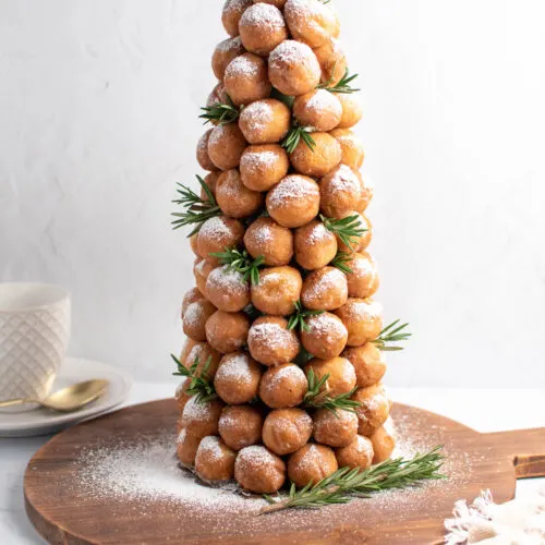 Donut hole tree with powdered sugar and fresh rosemary on wood cutting board with cup and saucer in background.