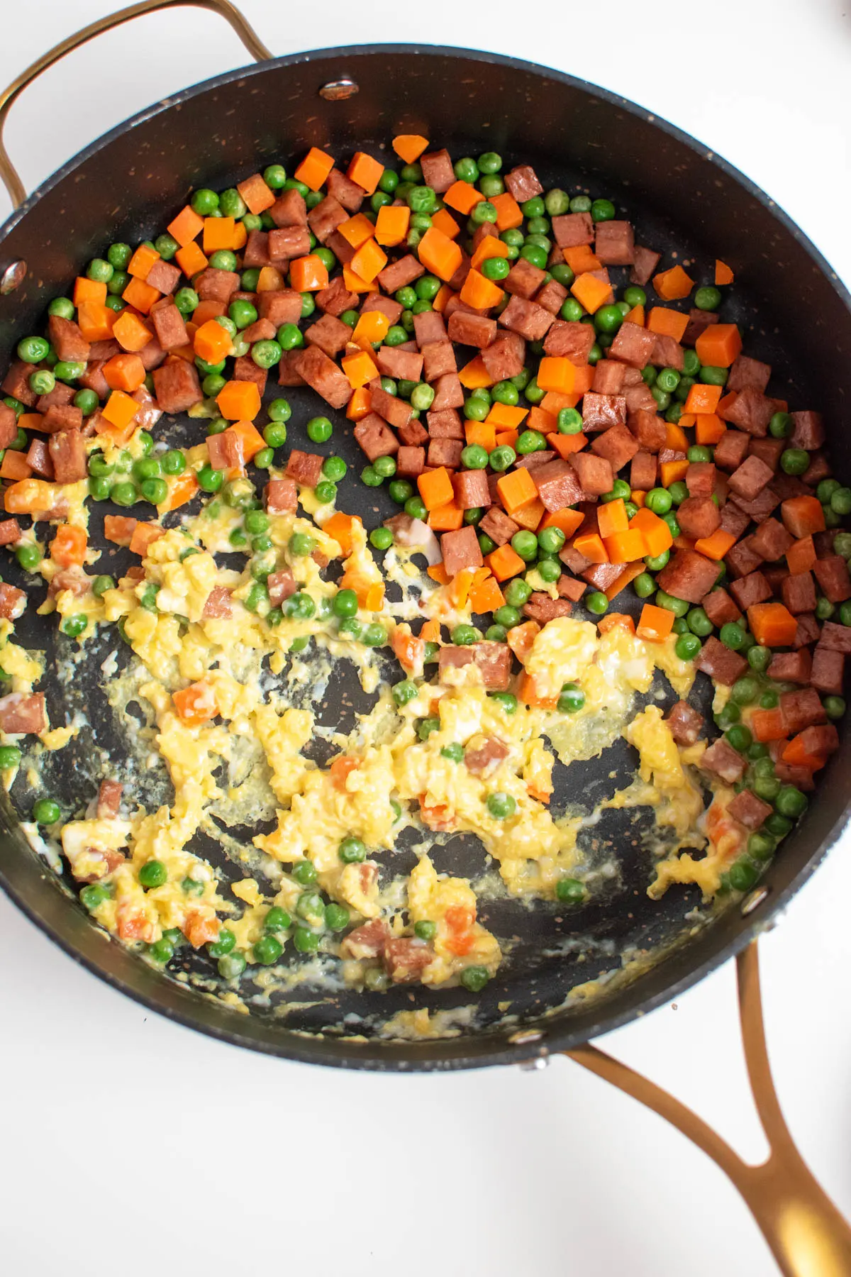 Partially cooked egg and small pieces of Spam, peas, and carrots in large black skillet.