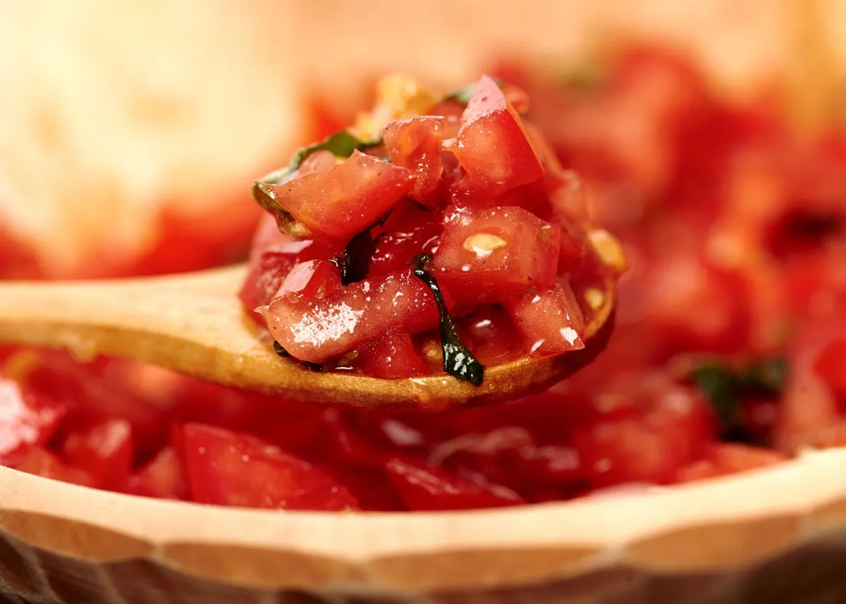 A wooden spoon lifts a mound of fire roasted diced tomatoes from a wooden bowl.