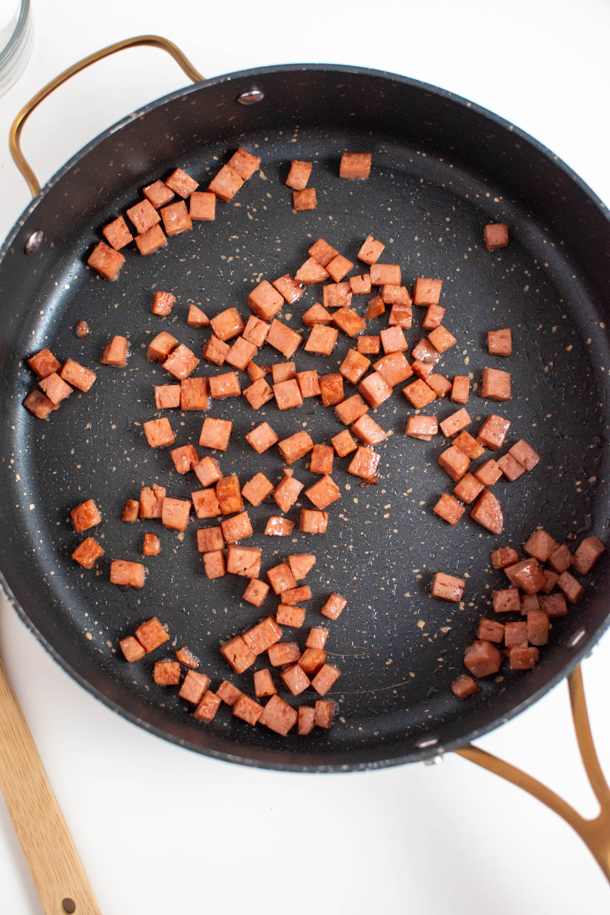 Several small fried Spam pieces in large black skillet on white table next to wood spoon.
