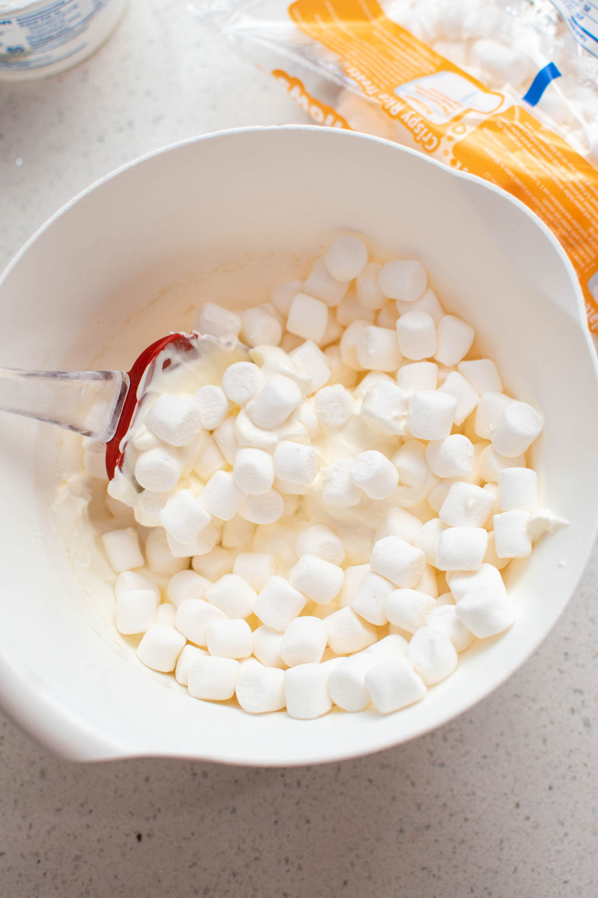 White bowl with mini marshmallows, sour cream, and spatula next to bag of mini marshmallows.