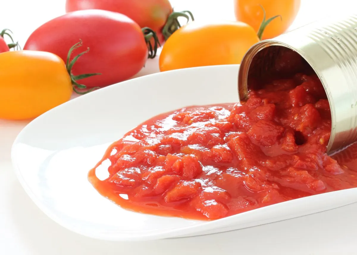 Petite diced tomatoes spilling from a can onto a white plate next to fresh tomatoes.