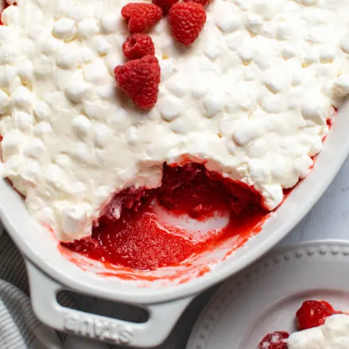 White baking dish with raspberry Jello salad and marshmallow topping with small piece removed.