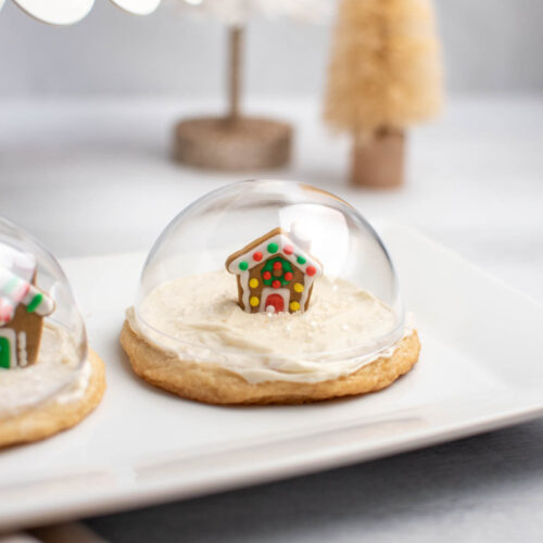 Snow globe sugar cookie with gingerbread house on white platter with tree decorations in background.