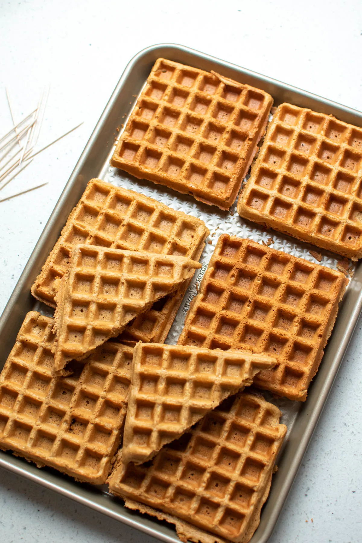 Large metal baking sheet with several gingerbread waffles and toothpicks, all on countertop.