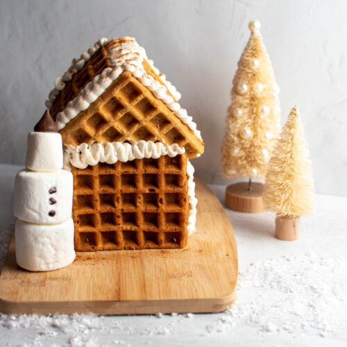Waffle gingerbread house with whipped cream and marshmallow snowman on wood cutting board with bottle brush trees nearby.
