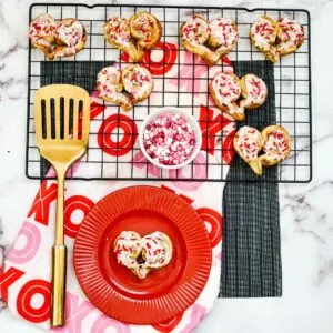 Heart-shaped cinnamon rolls on a baking rack with white glaze and pink and red sprinkles.
