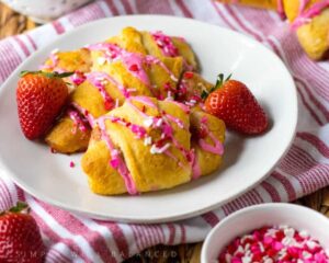 Two croissants on a white plate with pink frosting, sprinkles, and strawberries.