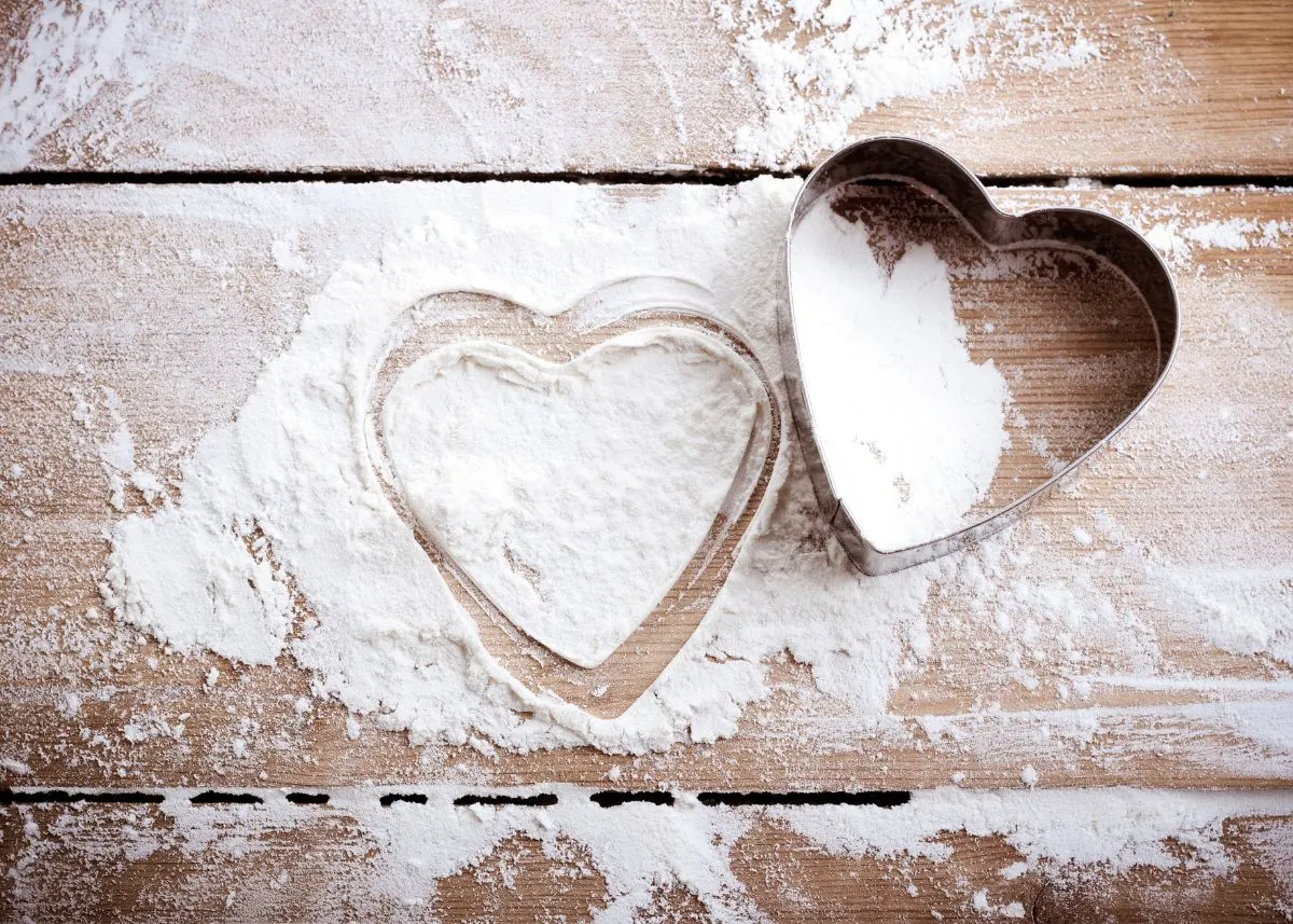 Two silver heart-shaped cookie cutters on a pile of flour on a wooden kitchen table.
