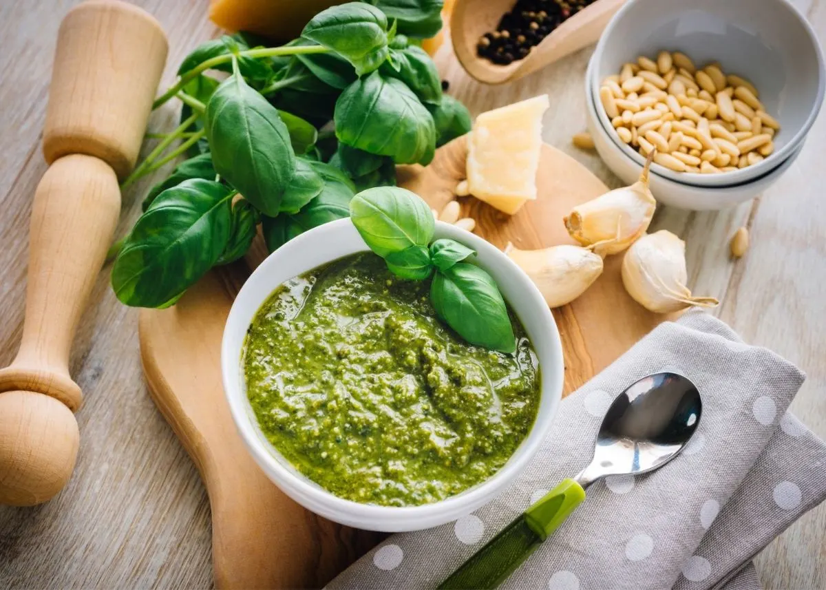 Pesto sauce in white bowl surrounded by garlic, parmesan, basil and pine nuts.