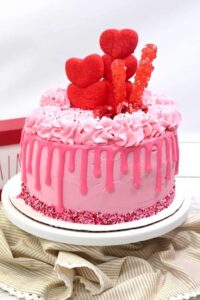 Pink Valentine's Day cake with drip and heart decorations on white platter.