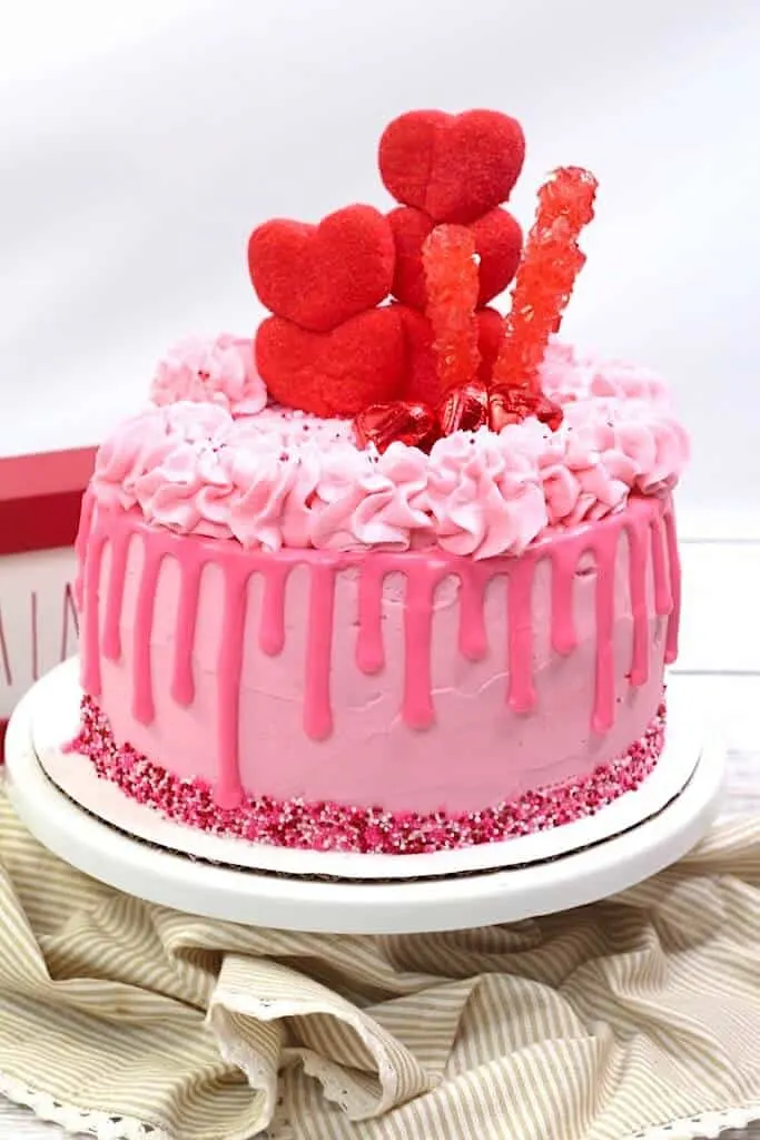 Pink Valentine's Day cake with drip and heart decorations on white platter.