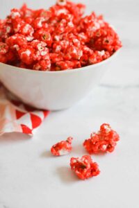Red candied popcorn in a white bowl next to a kitchen towel with hearts.