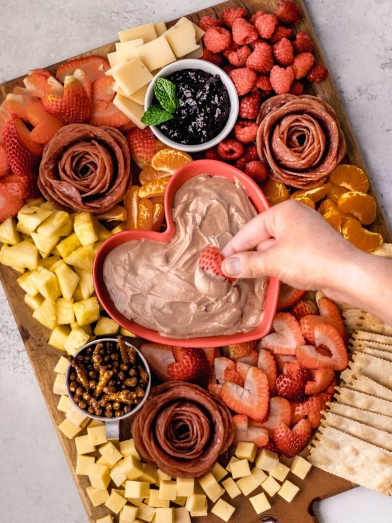 Hand dips strawberry in chocolate dip in heart shaped dish on charcuterie board.