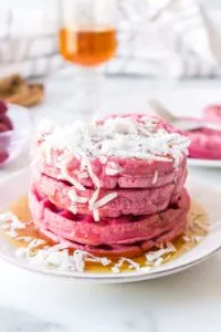 Stack of pink waffles with coconut shreds and syrup on a white plate.
