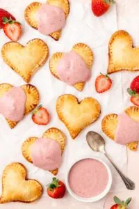 Heart-shaped strawberry hand pies with pink glaze next to fresh strawberries.