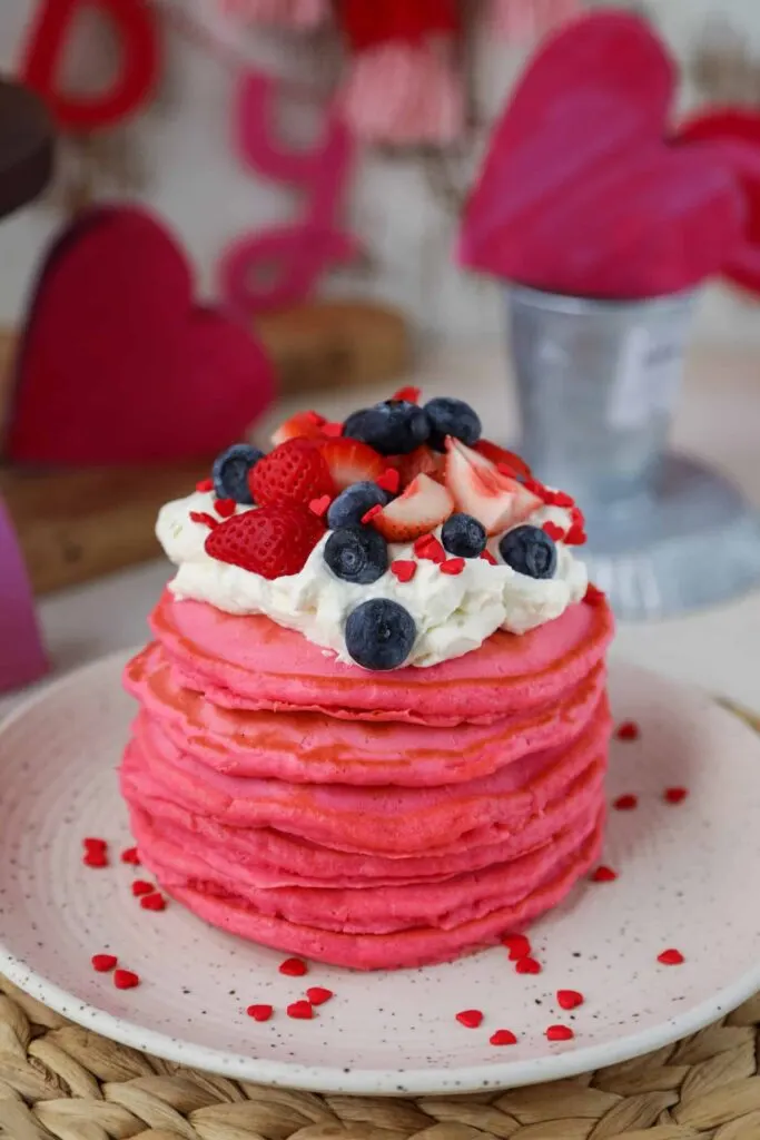 Tall stack of pink pancakes with fresh blueberries and strawberries on white plate.