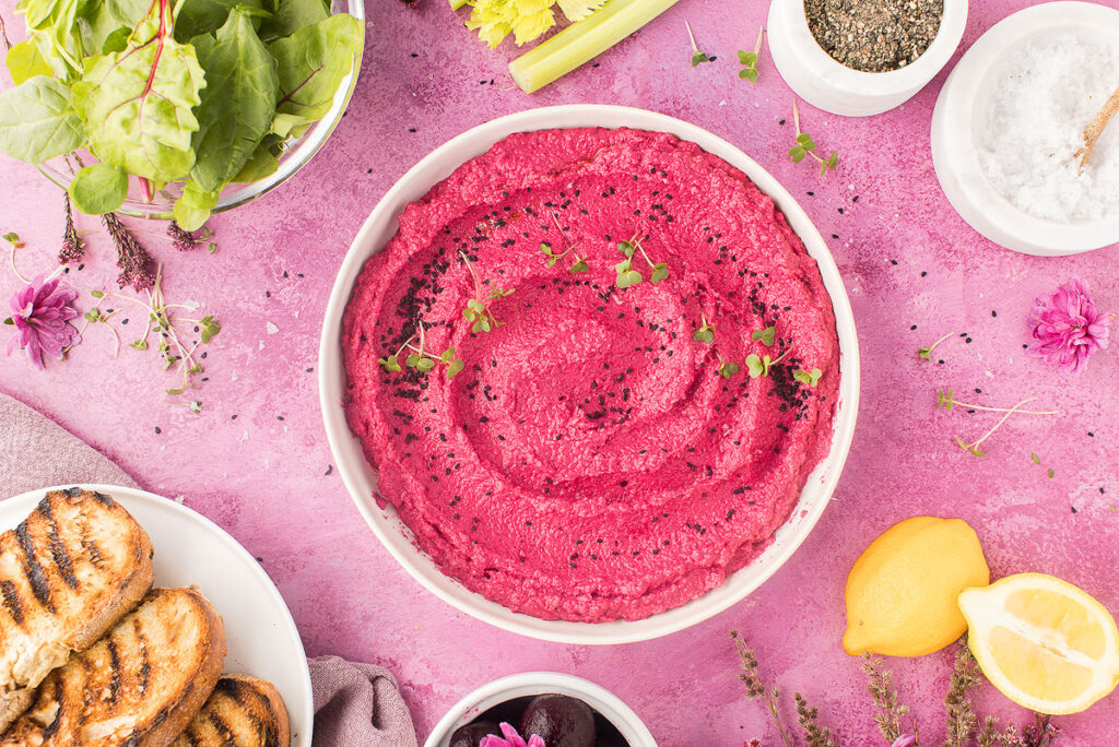 Pink beet hummus garnished with seeds on pink tablecloth with fresh ingredients surrounding.