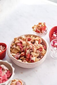 Sweet Chex mix in a white bowl with red and pink cookies and candies.