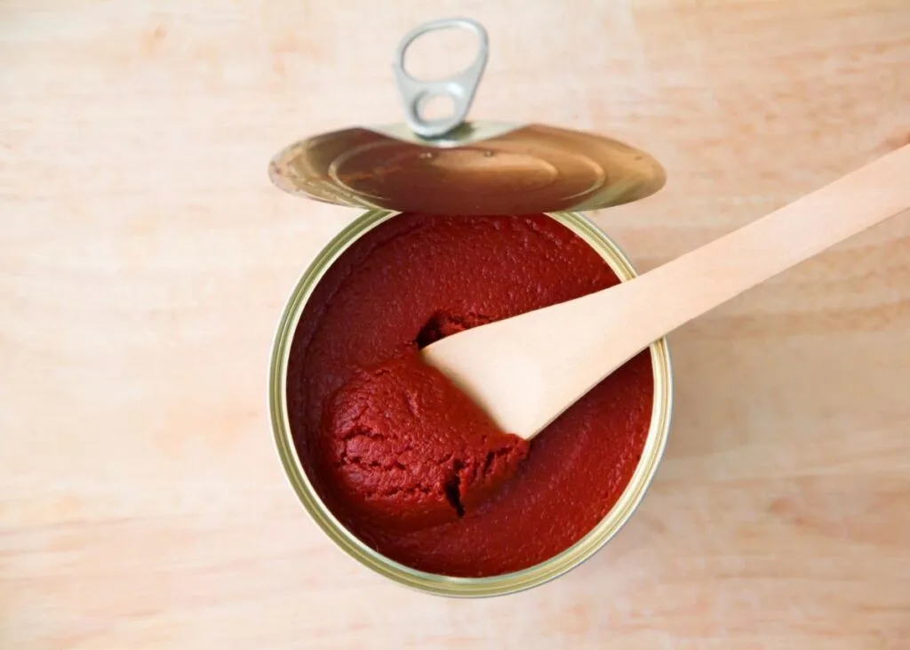Opened can of tomato paste with lid sticking out and wooden spoon inside.
