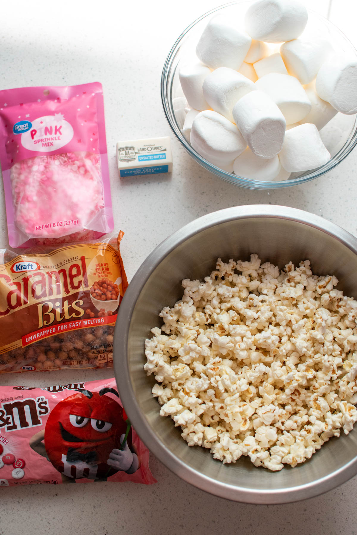 Ingredients for caramel popcorn cake on quartz counter including popcorn, marshmallows, and candy.