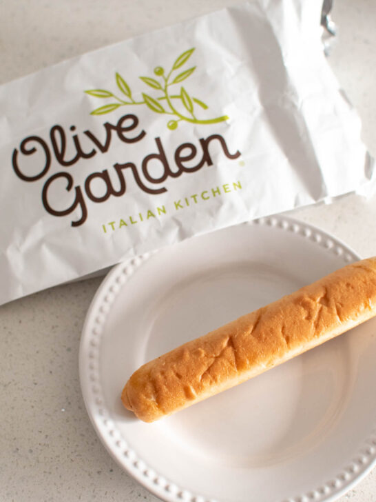 An Olive Garden breadstick on a plate next to the restaurant bag.