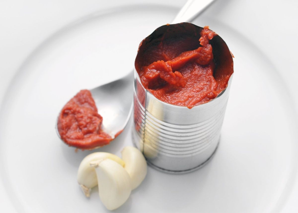 Tomato paste in can next to garlic cloves and spoon of paste on white plate.