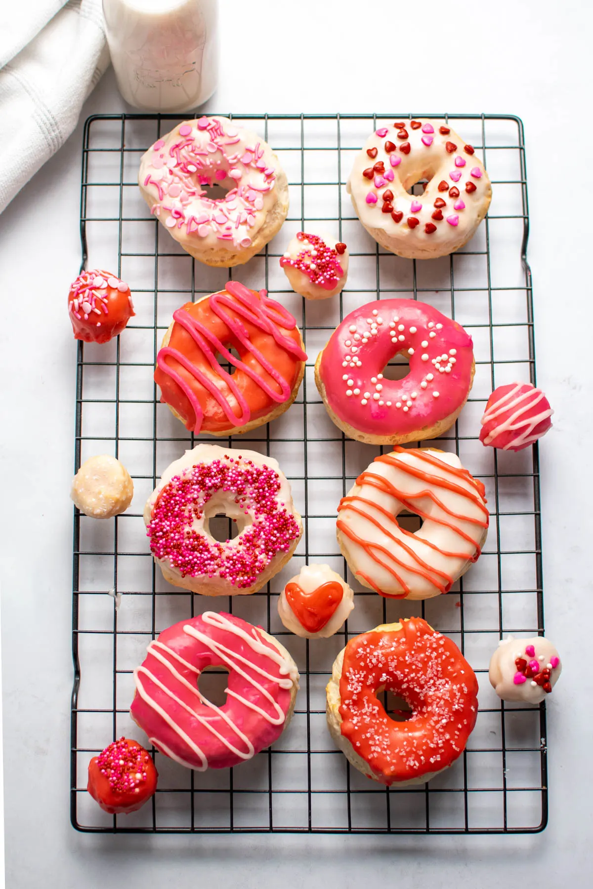 Several decorated Valentine's Day donuts and donut holes on black baking rack that's sitting on quartz countertop.