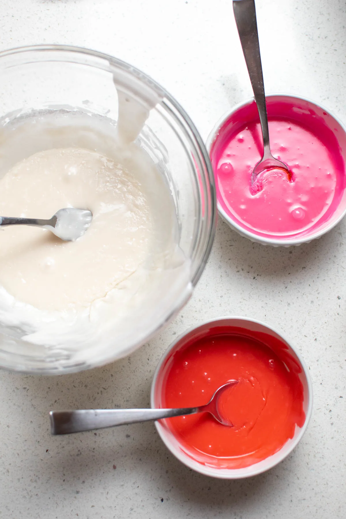 Bowls of white, pink, and red powdered sugar glaze and spoons on quartz counter.