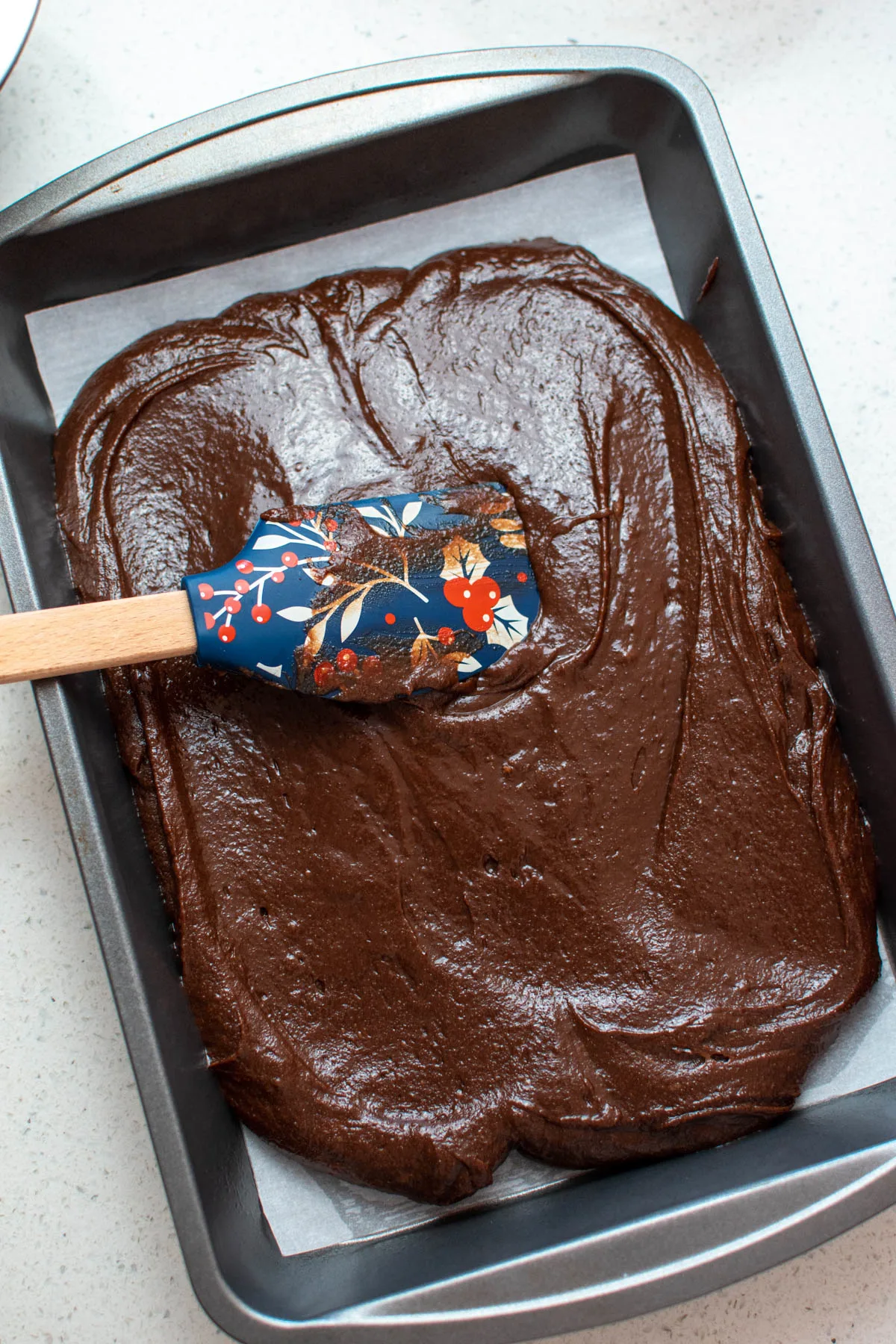 Rubber spatula rests in brownie batter in baking pan with parchment paper.