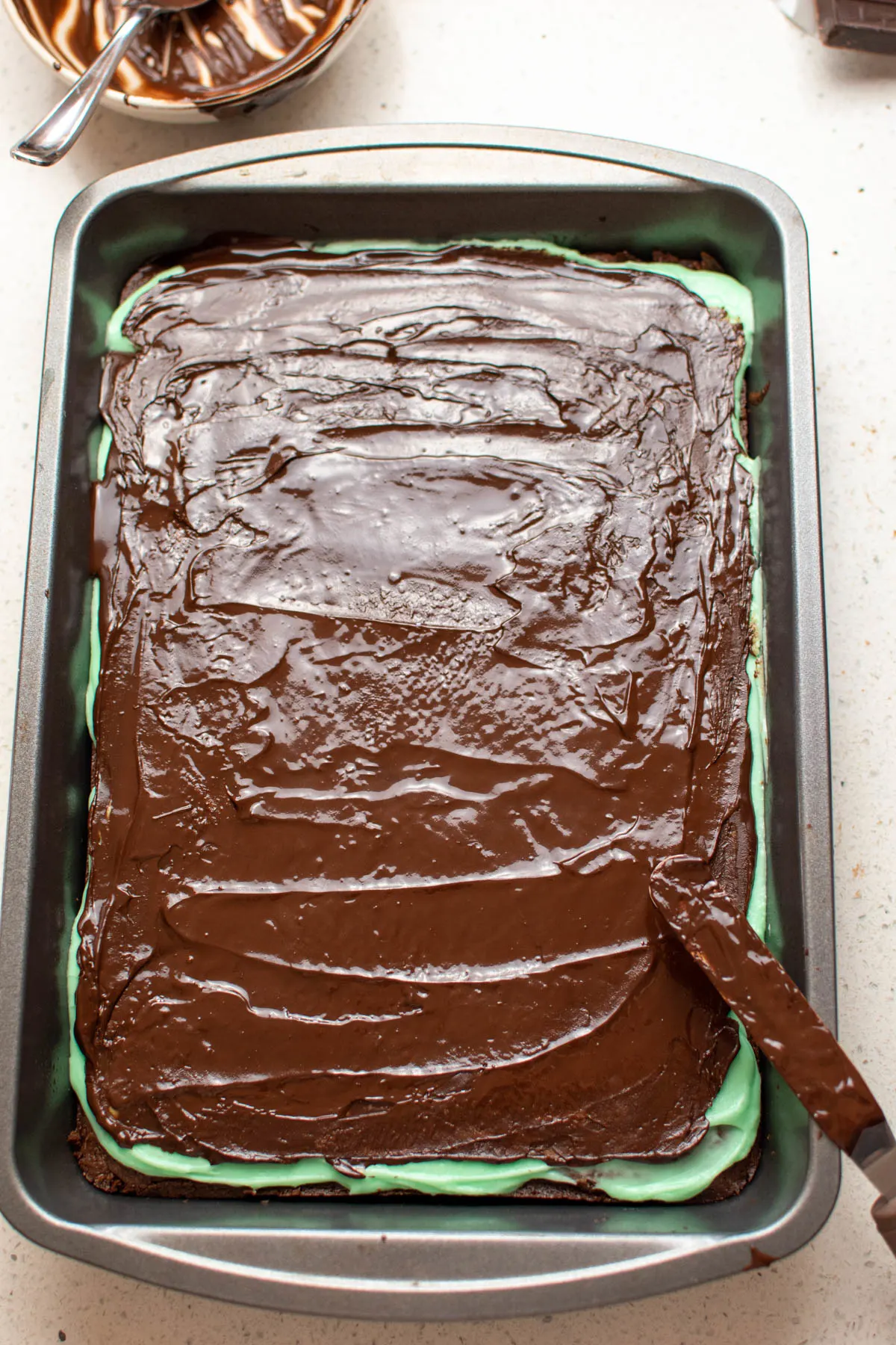 Chocolate ganache spread on pan of mint brownies sitting on quartz countertop.