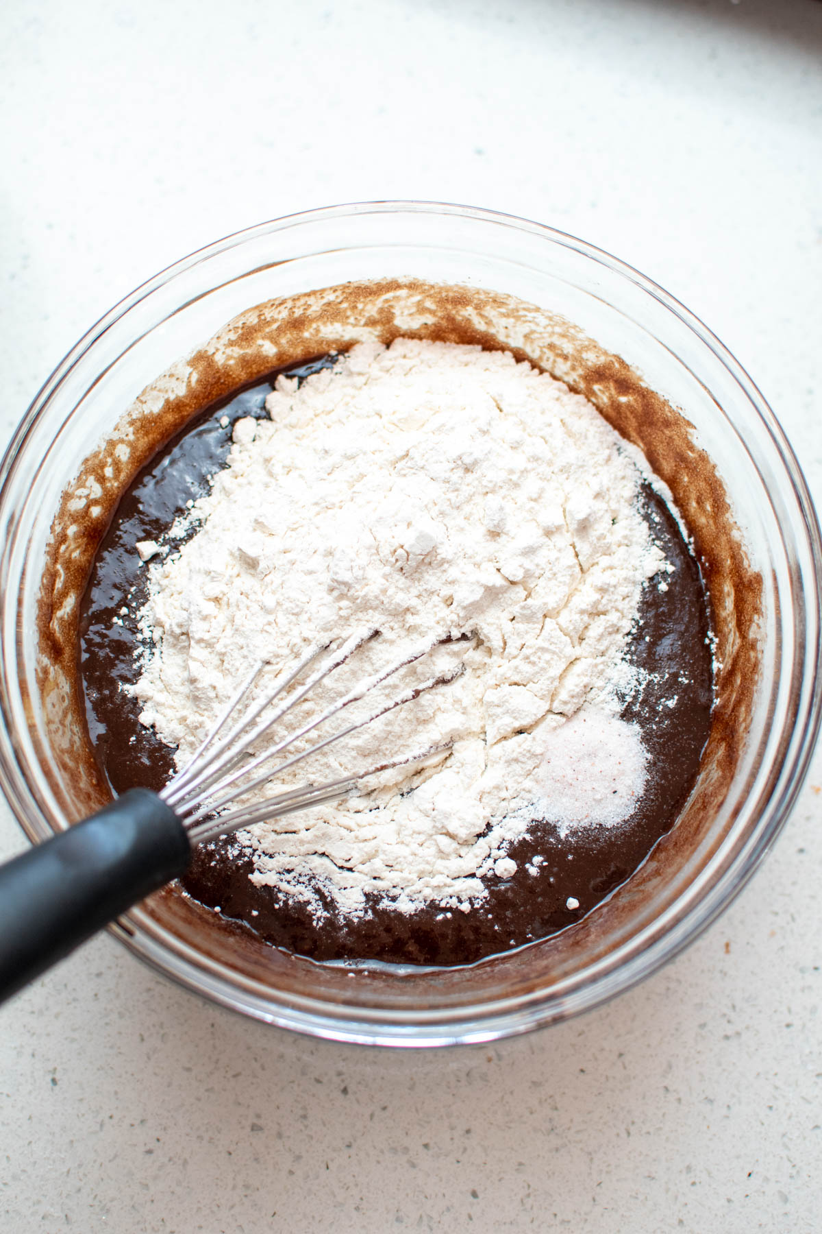 Flour and whisk in glass bowl of chocolate mixture on quartz countertop.