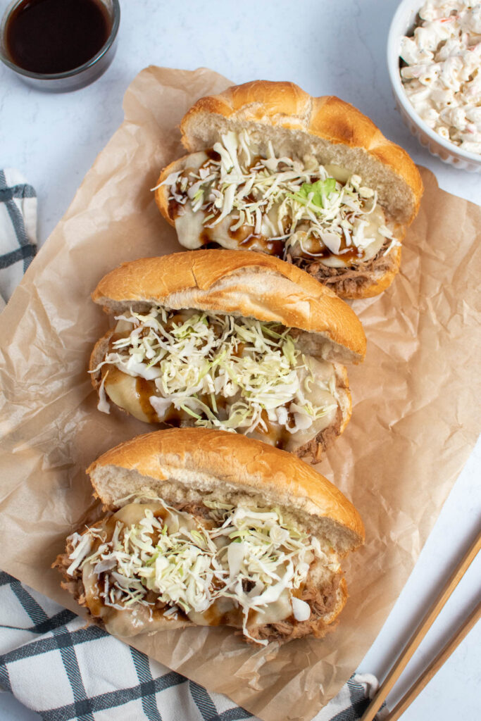 Three kalua pig sandwiches with shredded cabbage on brown parchment paper.