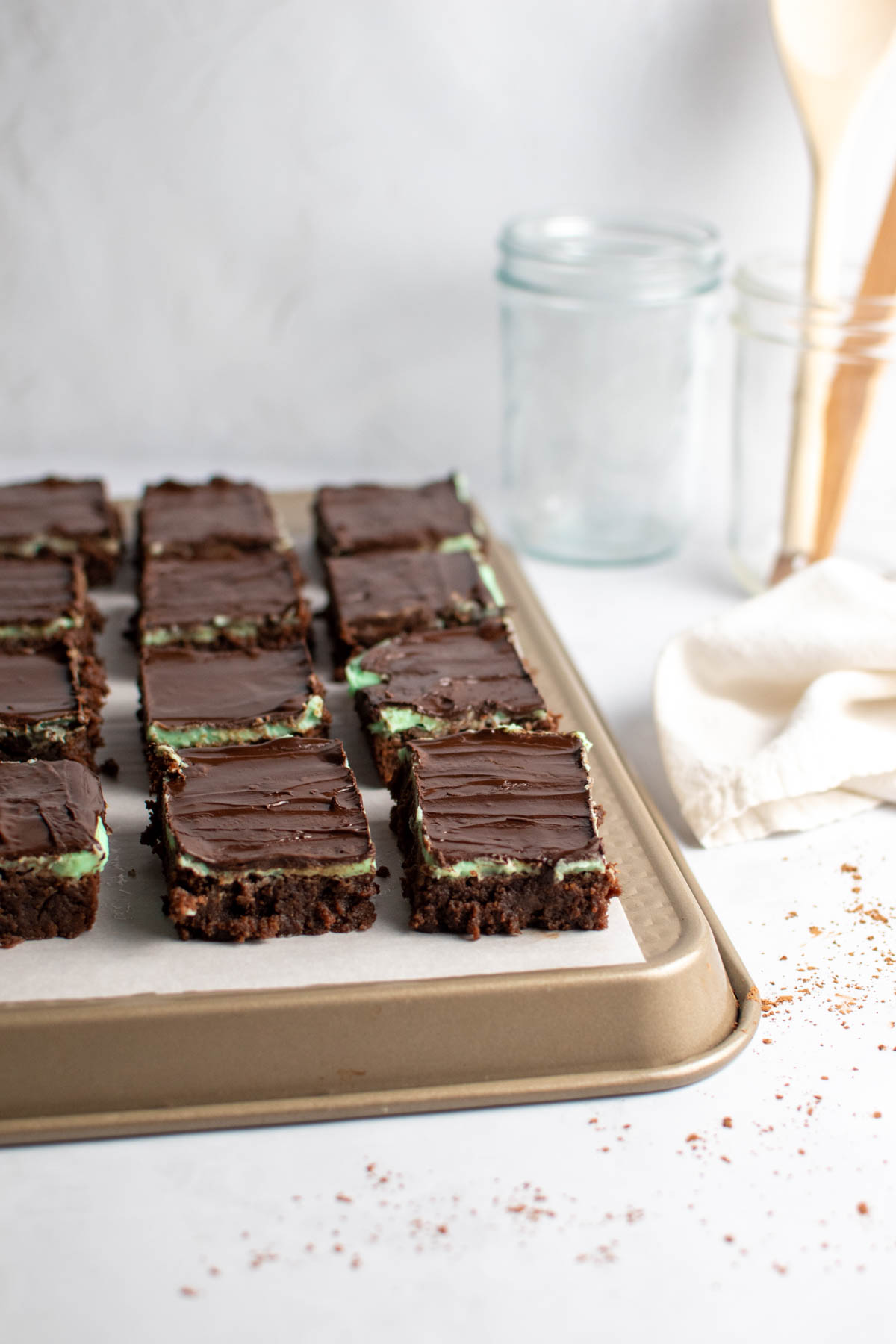 Several mint chocolate brownies on piece of parchment paper on baking sheet with utensils in background.