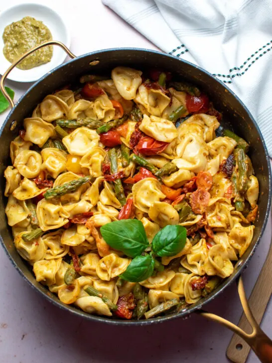 Pesto tortellini with tomatoes and asparagus in large black skillet on table top.