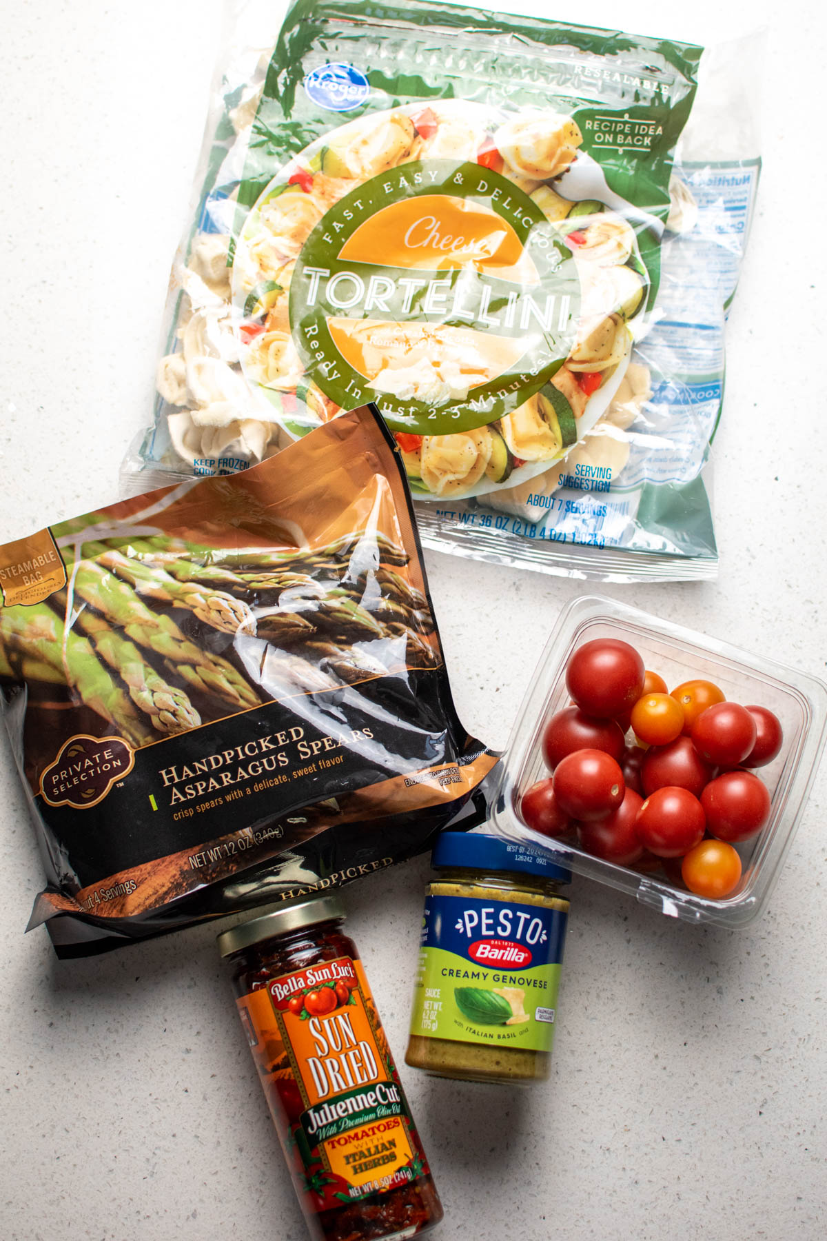 Pesto tortellini ingredients on quartz countertop including basil pesto, fresh tomatoes, and asparagus.