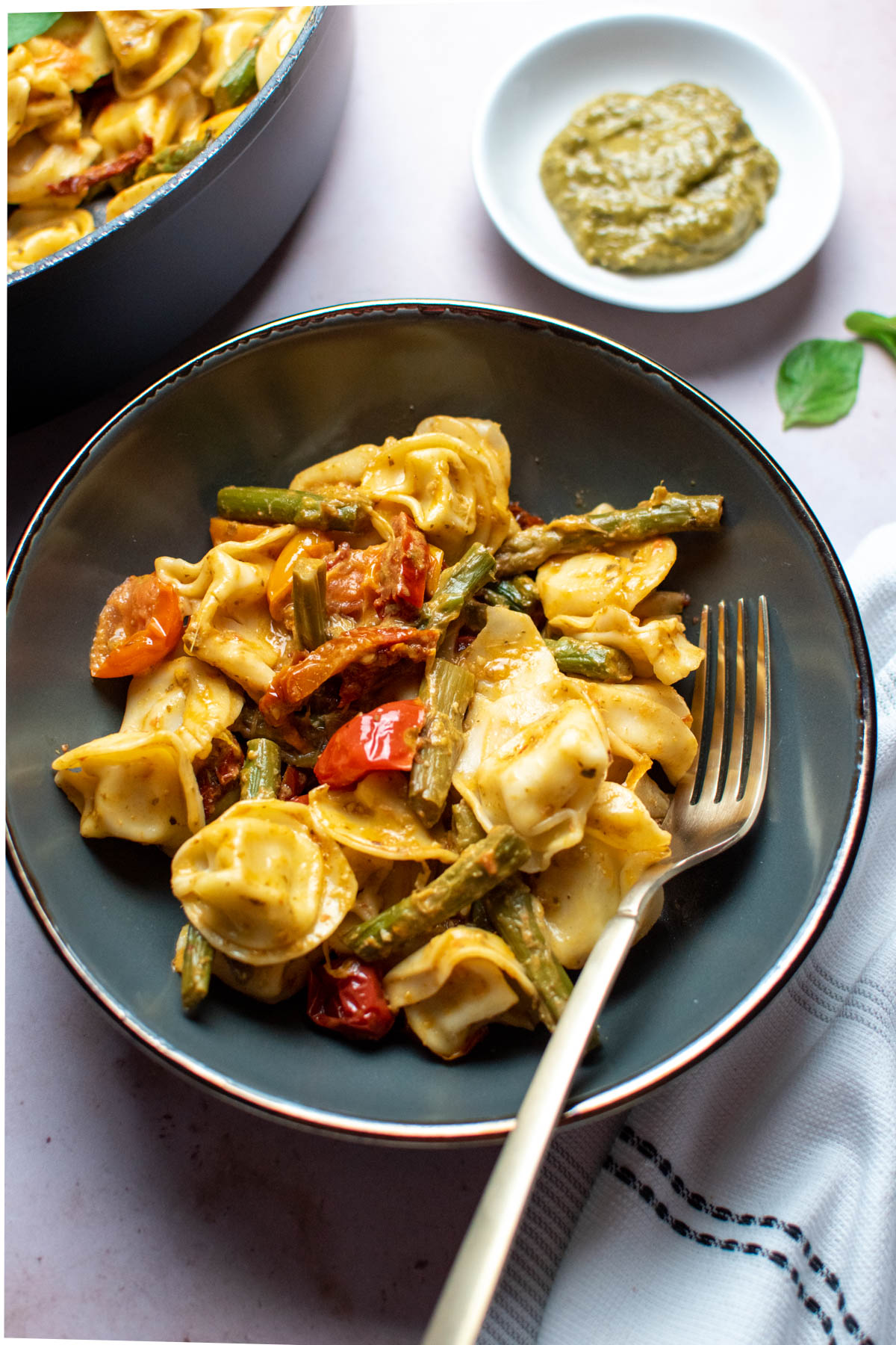 Pesto with tortellini and vegetables in blue gray bowl with gold fork.