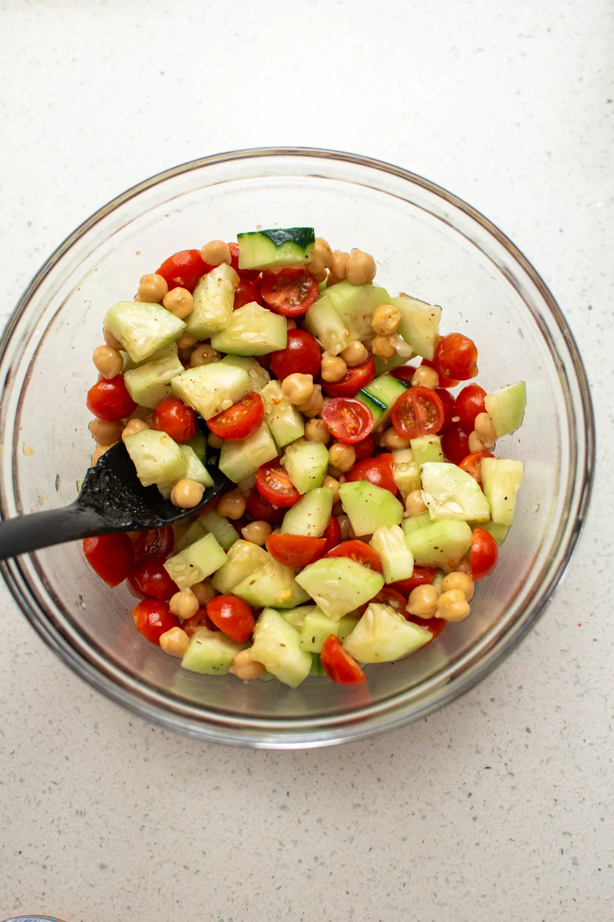 Tomato, cucumber, and chickpea salad in glass bowl with black spoon.