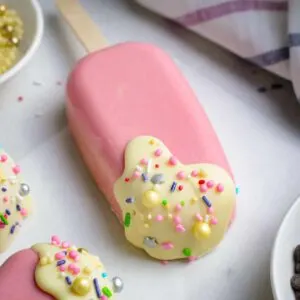 Pink brown cakesicle on a stick with white frosting and sprinkles.